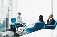 View through open glass door into a glass meeting room, four people sitting around a coffee table sitting on blue seats